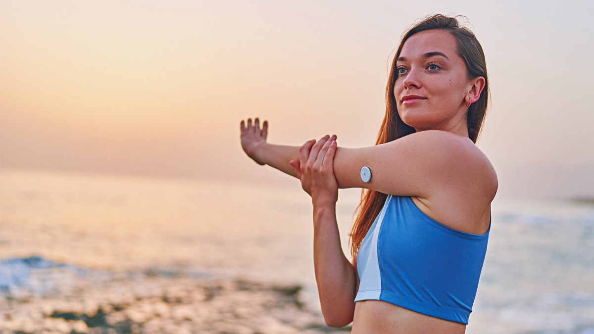 Satisfied One Fitness Slim Diabetic Patient Woman Doing Hands Stretching Exercises By The Sea. Healthy Habits And Active Sugar Diabetes Lifestyle A Woman Stretching Her Shoulder And Arm With A Glucose Monitor On Her Tricep.