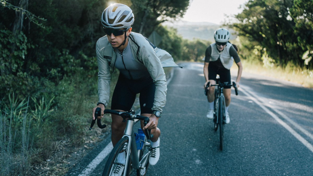 Two Road Cyclists Riding On Road Outside With Trees In The Background On Bikes
